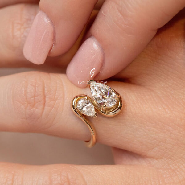 Close-up view of a yellow gold bypass ring worn on a finger, featuring two pear-shaped diamonds in an elegant asymmetrical design. One larger pear-cut diamond curves opposite a smaller matching stone, both secured in polished gold prongs. The smooth gold band wraps gently around the finger, creating a fluid, modern silhouette. Soft natural light enhances the sparkle of the diamonds and highlights the refined craftsmanship and romantic, contemporary jewelry style.