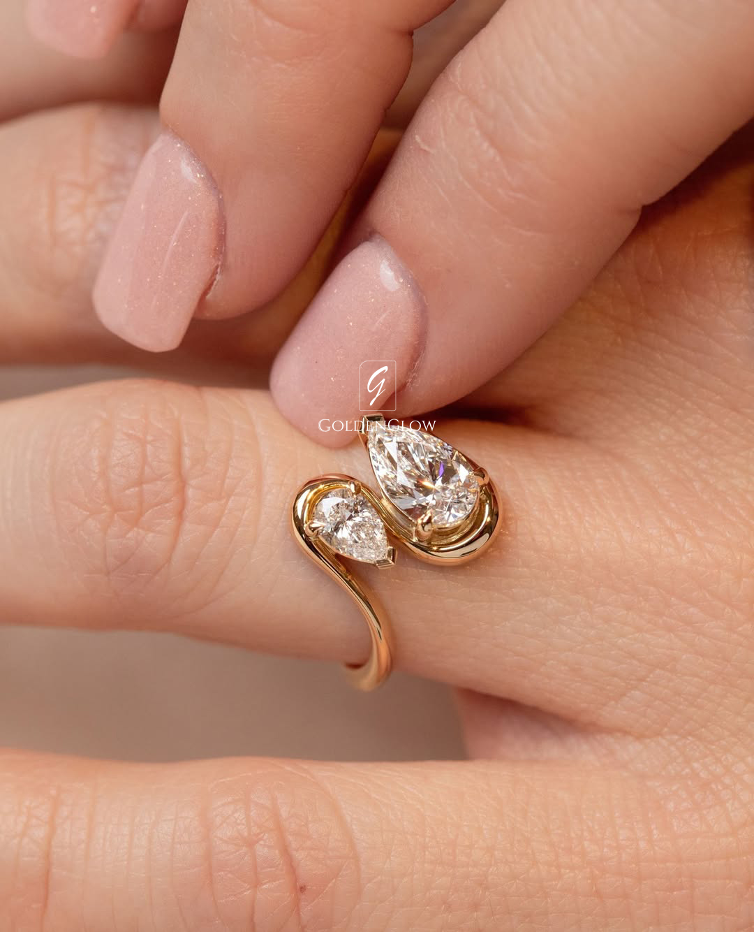 Close-up view of a yellow gold bypass ring worn on a finger, featuring two pear-shaped diamonds in an elegant asymmetrical design. One larger pear-cut diamond curves opposite a smaller matching stone, both secured in polished gold prongs. The smooth gold band wraps gently around the finger, creating a fluid, modern silhouette. Soft natural light enhances the sparkle of the diamonds and highlights the refined craftsmanship and romantic, contemporary jewelry style.