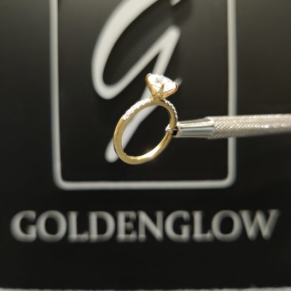Side view of a yellow gold engagement ring held by a jeweler’s tool, showcasing a marquise-cut diamond set high in a prong setting. The slim gold band features a subtle line of small accent diamonds along the top. The ring is photographed against a dark background with a softly blurred GoldenGlow logo, emphasizing the refined profile, sparkle, and craftsmanship.