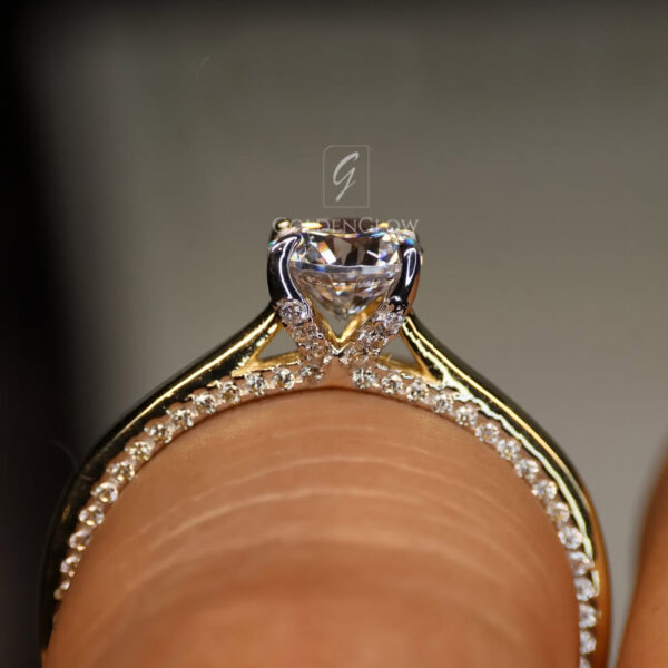 Macro side view of a yellow gold engagement ring worn on a finger, highlighting the under-gallery and setting. A round brilliant-cut diamond is held in a prong setting, elevated above a slim band adorned with pavé diamonds along the shoulders. The polished gold band curves smoothly around the finger. Fine skin texture is visible beneath the ring, while the background is softly blurred, drawing focus to the craftsmanship and diamond sparkle.