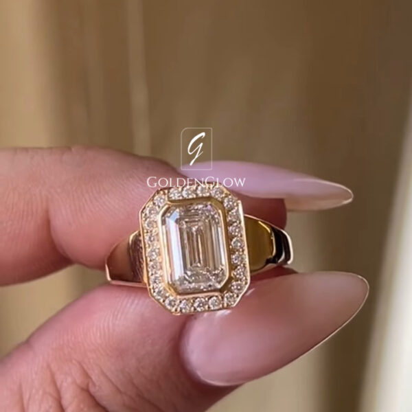 Close-up of a yellow gold ring held between fingers with long, manicured nails. The ring features an emerald-cut diamond set vertically at the center, surrounded by a rectangular halo of small pavé diamonds. The wide, polished gold band tapers slightly toward the setting. A soft, warm-toned background and subtle “GOLDENGLOW” watermark keep focus on the ring’s bold, elegant design and diamond clarity.