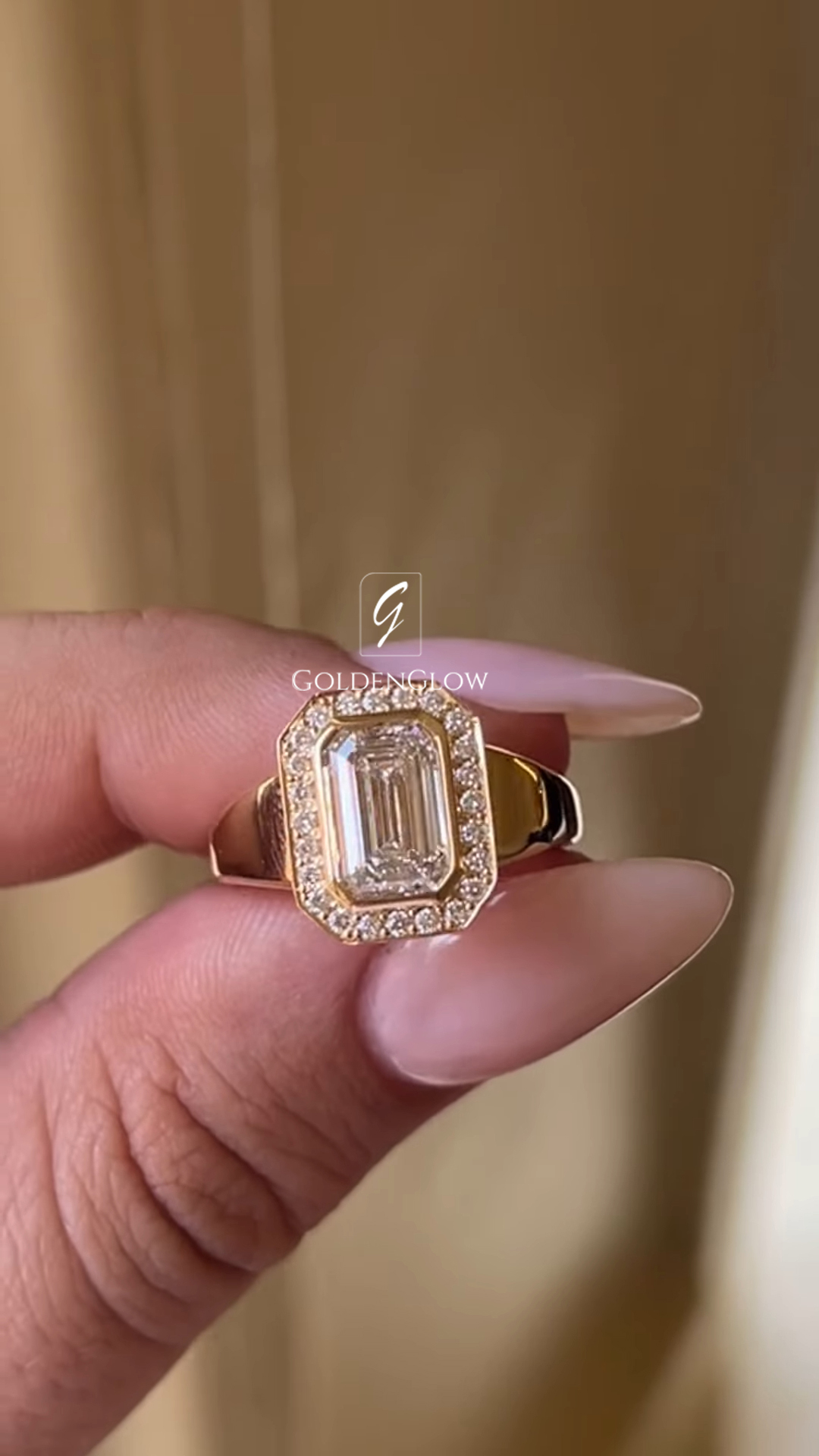 Close-up of a yellow gold ring held between fingers with long, manicured nails. The ring features an emerald-cut diamond set vertically at the center, surrounded by a rectangular halo of small pavรฉ diamonds. The wide, polished gold band tapers slightly toward the setting. A soft, warm-toned background and subtle โGOLDENGLOWโ watermark keep focus on the ringโs bold, elegant design and diamond clarity.