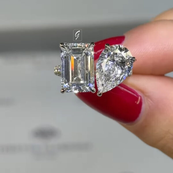 Close-up comparison image of two diamonds held between fingers with glossy red nail polish. On the left is an emerald-cut diamond in a four-prong setting, showing long step-cut facets and a clean rectangular shape. On the right is an oval-cut diamond in a prong setting, displaying rounded edges and brilliant facets with strong sparkle. The background is softly blurred in light gray tones, with a subtle GOLDENGLOW watermark centered above the stones.