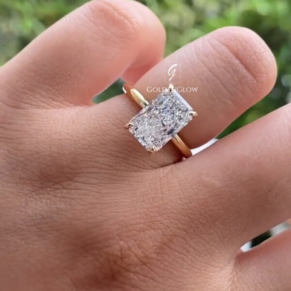 A close-up view of a hand wearing a yellow gold ring featuring a rectangular, emerald-cut lab-grown diamond secured with four slim prongs at each corner. The polished gold band is smooth and minimal, drawing attention to the diamond’s clean lines and step-cut facets. The center stone reflects natural light with sharp flashes and clarity. A softly blurred green outdoor background provides contrast, highlighting the ring’s elegant simplicity and , modern design.