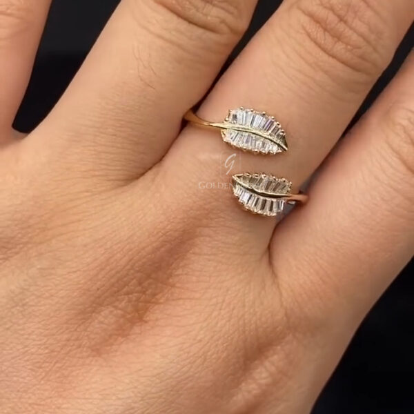 A close-up view of a hand wearing a yellow gold open-style ring with two leaf-shaped ends facing each other. Each leaf is decorated with rows of slim baguette-cut diamonds arranged in a vein-like pattern, creating structured sparkle and texture. The polished gold band curves smoothly around the finger in a modern bypass design. The diamonds reflect light with subtle brilliance, while the dark, softly blurred background enhances contrast and highlights the ring’s elegant, nature-inspired form.Art