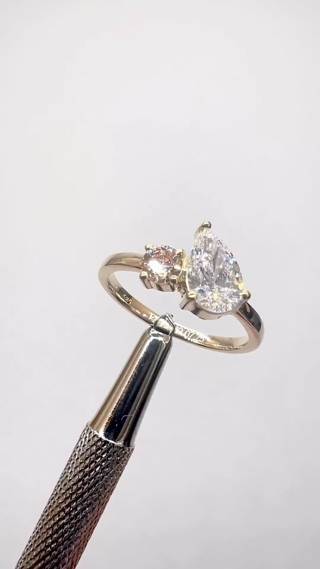 A close-up studio image of a yellow gold ring held by a jeweler’s tool against a white background. The ring features a pear-shaped brilliant-cut diamond set in prongs, paired with a smaller round accent diamond beside it in an asymmetrical design. The polished gold band curves smoothly and reflects light, while both diamonds show bright sparkle and clarity, emphasizing the ring’s modern and elegant craftsmanship.