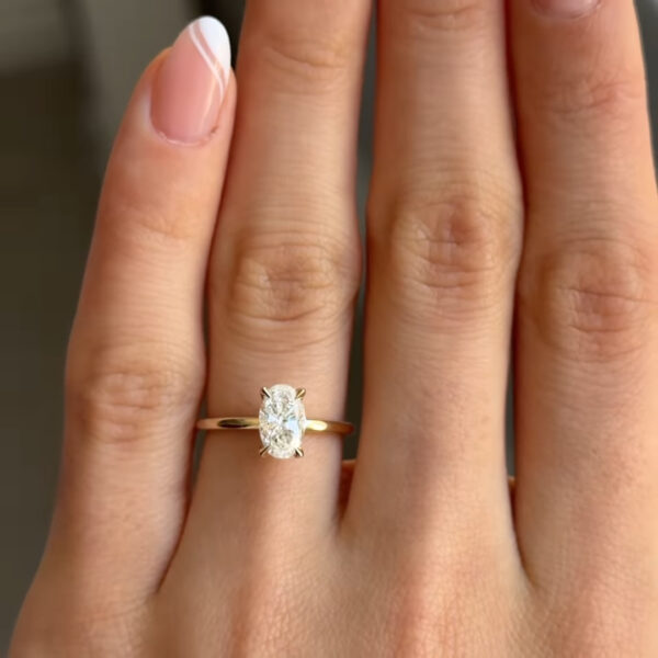 A close-up view of a hand wearing a yellow gold ring with an oval brilliant-cut diamond set vertically in a four-prong setting. The slim, polished gold band sits smoothly on the finger, allowing the elongated diamond to remain the focal point. The diamond reflects soft light with clear sparkle and defined facets, while the natural skin tones and neutral background emphasize the ring’s clean, elegant, and design.