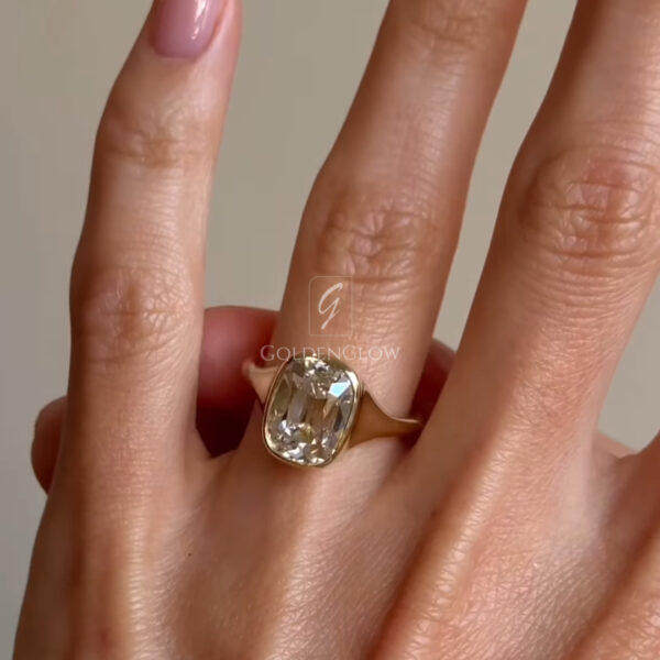 Close-up of a hand holding a solitaire ring between the thumb and index finger. The ring features an elongated cushion-cut clear gemstone set in a smooth yellow gold bezel-style band. The stone is available in both lab-grown diamond and moissanite options, each offering bright sparkle and sharp, reflective facets. Soft natural light highlights the gemstone’s clarity and depth. The fingers have a glossy pale pink manicure, and a neutral background keeps focus on the ring’s modern, elegant design.