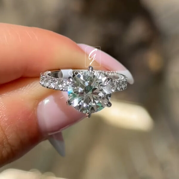 Close-up of a hand holding a diamond engagement ring between the thumb and index finger. The ring features a round brilliant-cut clear center stone secured by four prongs, set on a white metal band adorned with small pavé diamonds along the shoulders. The center stone reflects bright natural light with sharp, sparkling facets. The fingers have a pale, glossy manicure, and natural skin texture is visible.
