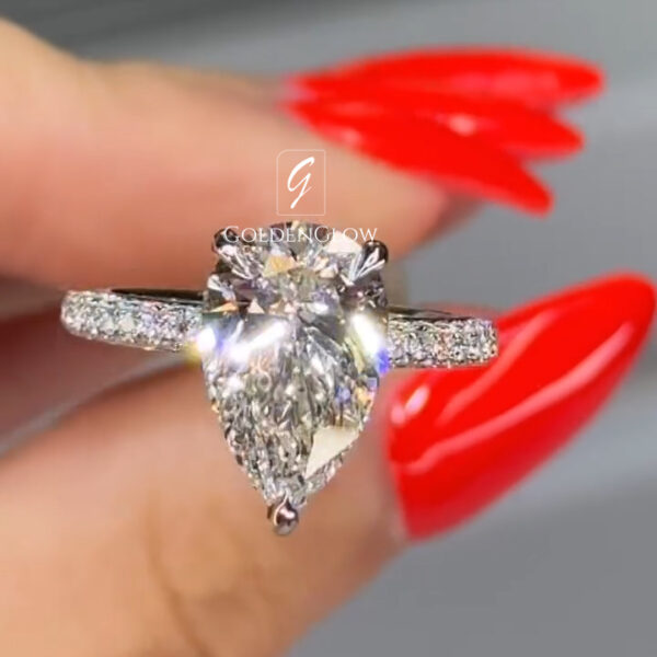 Close-up of a hand holding a diamond engagement ring between the thumb and index finger. The ring features a pear-shaped clear center stone secured by prongs, set on a white metal band with pavé diamonds along the shoulders. The center stone reflects bright light with sharp, sparkling facets. The fingers have long, glossy red nails that contrast with the silver-toned metal. A smooth gray background keeps focus on the ring’s brilliance, elegant silhouette, and detailed craftsmanship.