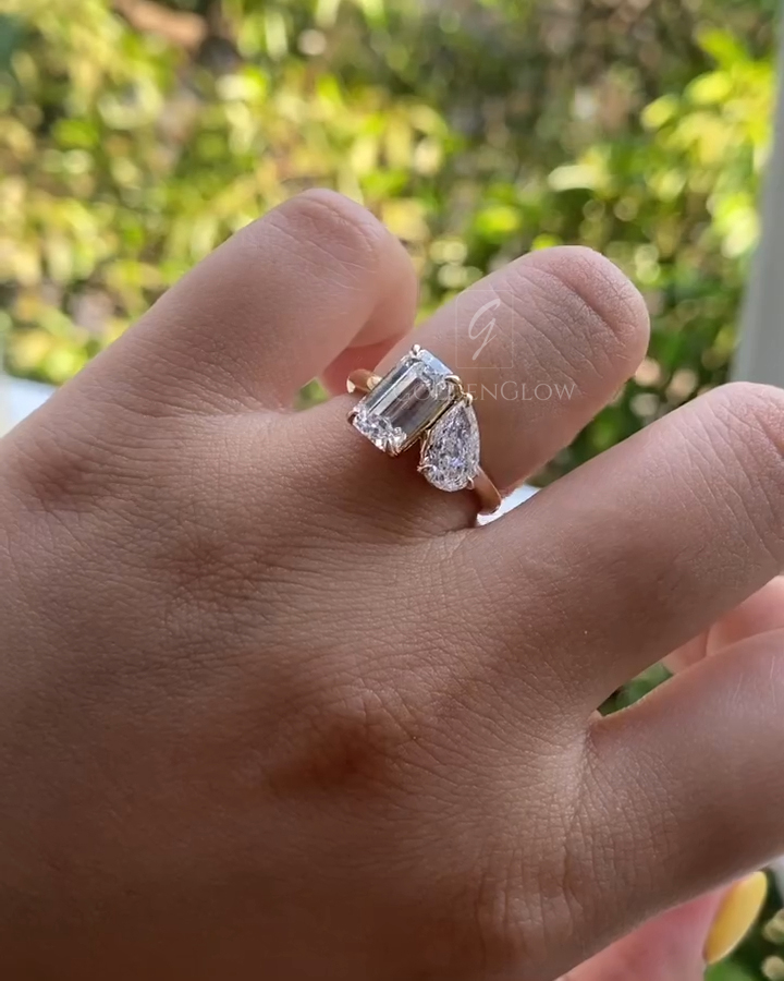 Hand wearing a toi et moi ring with an emerald-cut clear stone and a pear-cut clear stone set side by side on a slim yellow gold band. Natural daylight highlights the crisp step facets and elegant asymmetrical design against a leafy green background.