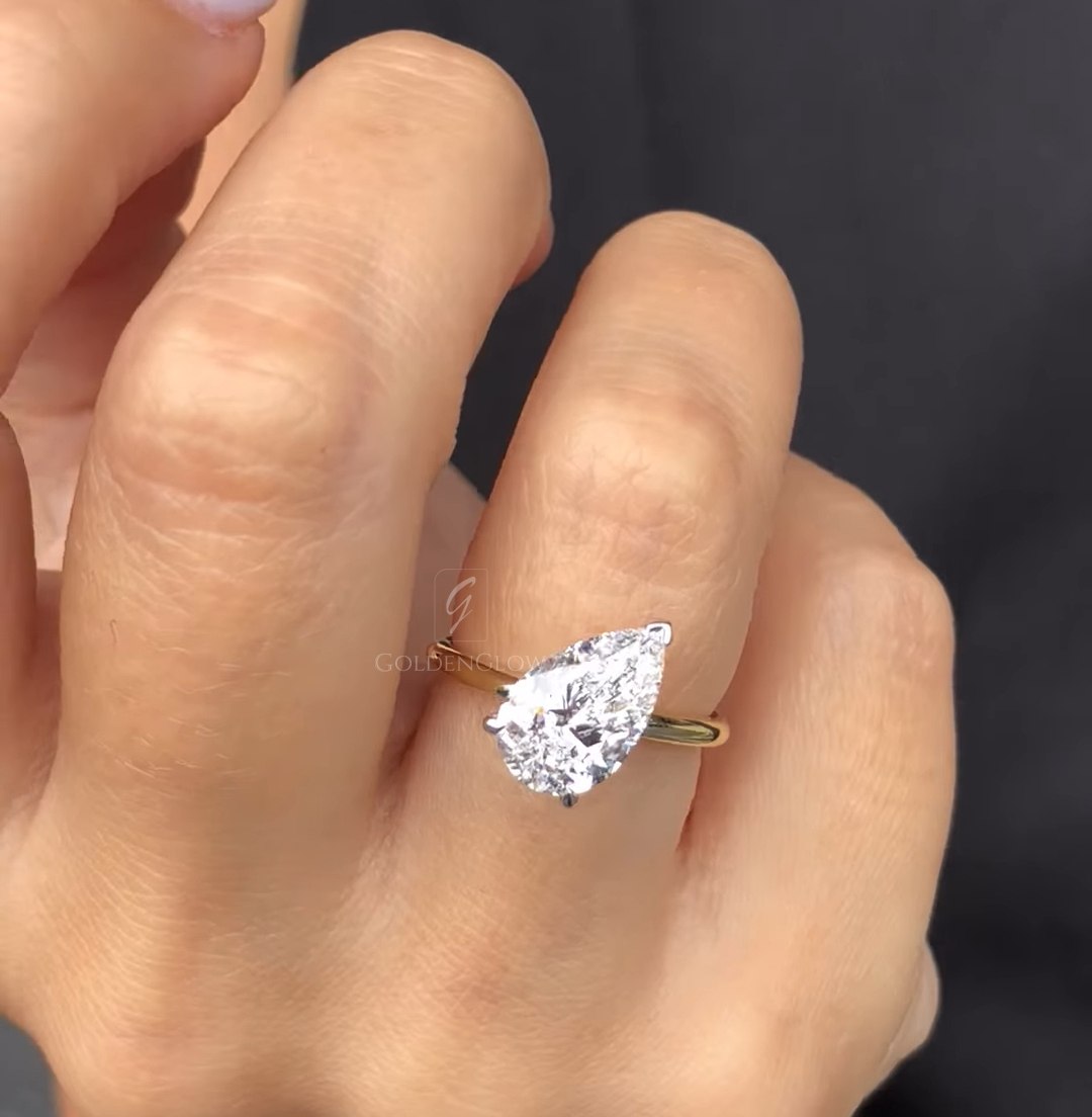 Close-up of a two-stone ring held between fingers, featuring an emerald-cut clear lab-grown diamond paired with a pear-shaped green gemstone. The stones are set side by side in polished white metal, highlighting clean lines, sharp facets, and a modern asymmetrical design against a soft neutral background.