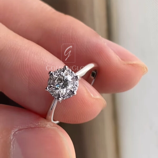 Close-up photograph of a round brilliant diamond engagement ring held between fingers in natural light. The center diamond is set in a white gold or platinum band with a delicate halo of small accent diamonds surrounding the main stone. The prong setting and polished band reflect light, highlighting the sparkle, clarity, and craftsmanship of the ring against a softly blurred background.