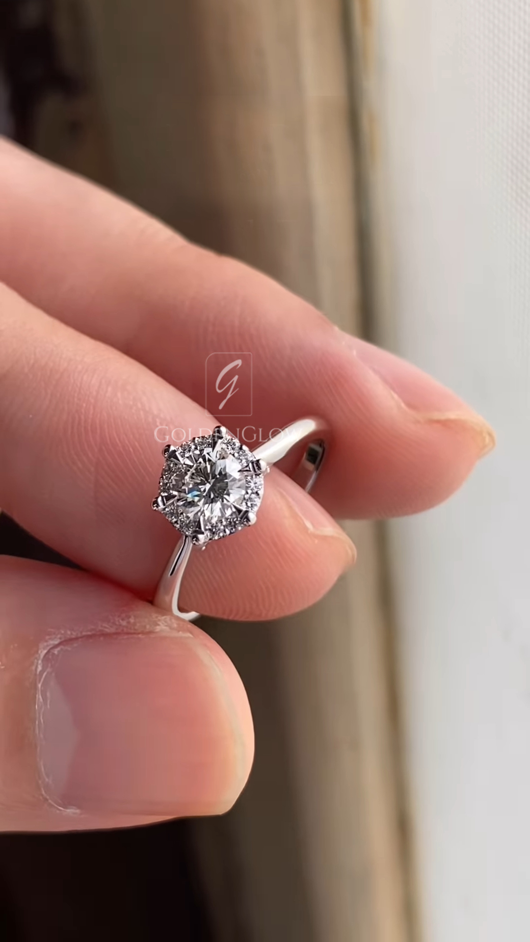 Close-up photograph of a round brilliant diamond engagement ring held between fingers in natural light. The center diamond is set in a white gold or platinum band with a delicate halo of small accent diamonds surrounding the main stone. The prong setting and polished band reflect light, highlighting the sparkle, clarity, and craftsmanship of the ring against a softly blurred background.