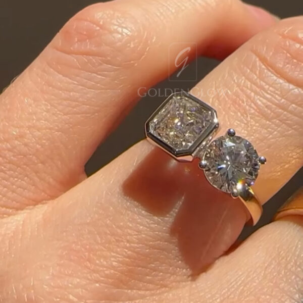 Detailed macro photograph of a toi et moi diamond ring shown from a slightly angled top view on a hand. The hexagon-shaped diamond displays step-cut facets within a geometric bezel, positioned next to a round brilliant diamond with visible prongs. Light reflections emphasize the contrast between the angular and circular stones. The polished band curves gently around the finger, with warm tones and soft shadows creating depth.