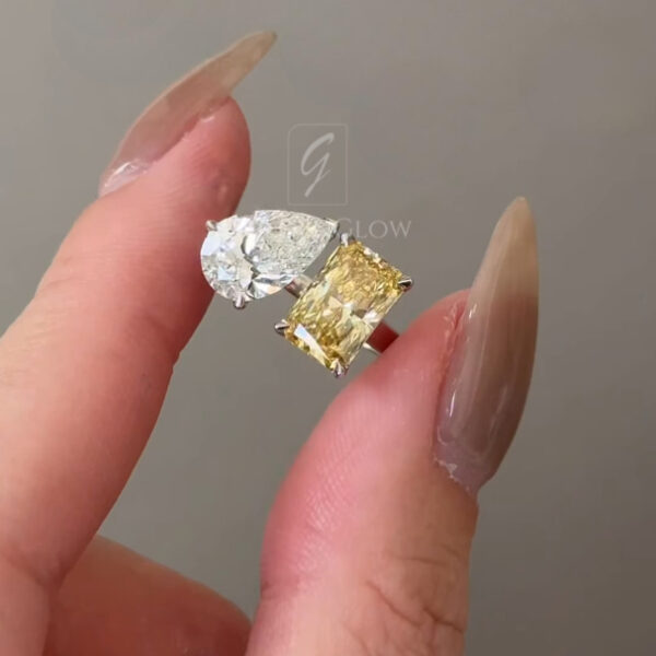 Close-up of an open bypass-style ring worn on a hand, featuring two gemstones set opposite each other. One stone is a pear-shaped clear gemstone in a white metal prong setting, and the other is a rectangular yellow gemstone set in a white metal prong mount. The polished band curves gracefully to frame both stones. Neutral background and natural lighting highlight the contrast between the clear and yellow stones.