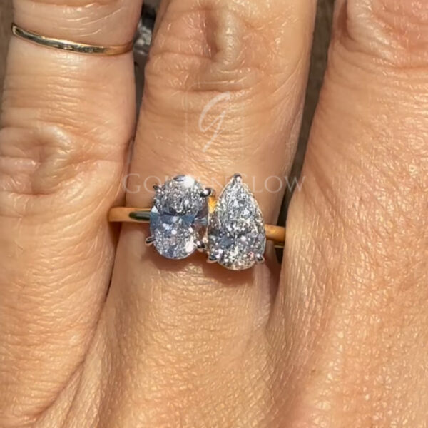 Close-up of a yellow gold toi et moi ring worn on a hand, featuring two sparkling diamonds—one oval cut and one pear cut—set side by side in a modern asymmetrical design against a rustic wooden background.