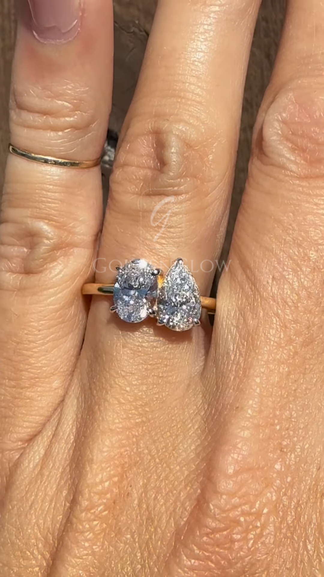 Close-up of a yellow gold toi et moi ring worn on a hand, featuring two sparkling diamonds—one oval cut and one pear cut—set side by side in a modern asymmetrical design against a rustic wooden background.