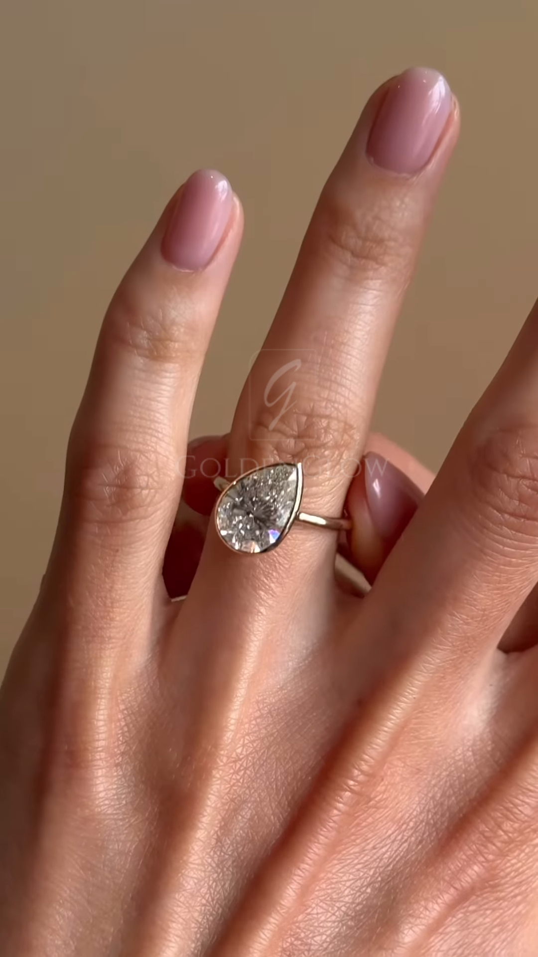 Close-up view of a pear-shaped diamond engagement ring worn on a hand with soft pink manicured nails. The teardrop-cut diamond is bezel set in polished yellow gold, catching light with bright facets. The ring sits naturally on the finger against a warm neutral background, highlighting the elegant silhouette, minimalist gold band, and refined sparkle suitable for an engagement or statement ring.