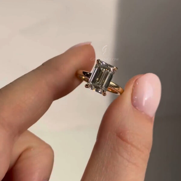Side angle view of an emerald-cut diamond ring pinched gently between thumb and finger, showcasing the vertical step facets and secure four-prong yellow gold setting. The polished gold band curves smoothly beneath the stone, while subtle reflections emphasize the diamond’s geometric brilliance and refined craftsmanship.