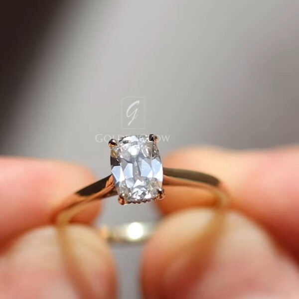 Side profile photograph of a handcrafted gold solitaire ring shown between fingers against a soft gray background. The radiant-cut diamond is elevated in an open basket setting, revealing the detailed prong structure beneath the stone. The slim gold band arches gracefully, showcasing a lightweight and comfortable design. The angle emphasizes the height of the setting and allows light to pass through the diamond, enhancing brilliance, transparency, and fine jewelry craftsmanship.