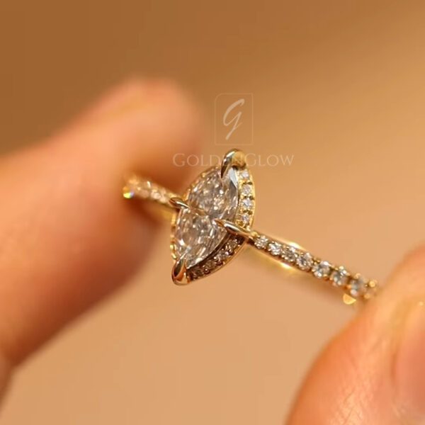 Detailed side-angle view of a marquise diamond ring crafted in polished yellow gold, shown between fingers in a softly lit studio setting. The elongated diamond is framed by a halo of small round diamonds, while the thin band is lined with pavé stones that extend along the shoulders. The warm lighting emphasizes the gold tone, intricate setting, and delicate profile of the ring, showcasing its refined proportions and graceful silhouette.