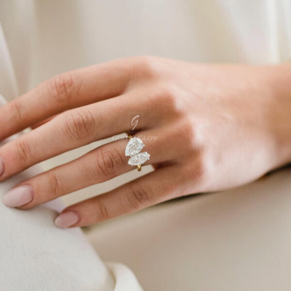 A close-up of a hand resting on soft white fabric, wearing a delicate engagement ring with two pear-cut moissanite or lab grown diamonds. The stones are arranged in a vertical, modern design on a slim yellow-gold band. The diamonds reflect gentle natural light, highlighting their brilliance and clarity. The hand features natural nude-pink manicured nails, and the light, neutral background enhances the elegant, minimal, and timeless appeal of the ring.