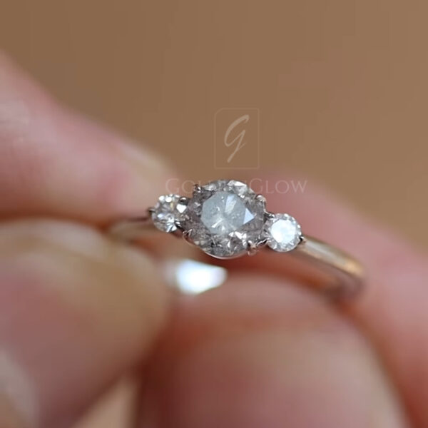 A detailed macro shot of a three-stone diamond ring resting across a finger, captured in soft natural lighting. The center round-cut diamond displays visible facets and subtle inclusions, framed by two smaller round diamonds on either side. The slim, polished metal band curves smoothly around the finger, reflecting light gently. The warm, neutral background and shallow focus draw attention to the gemstone brilliance, prong setting, and refined craftsmanship, creating a timeless and intimate jewelry presentation.
