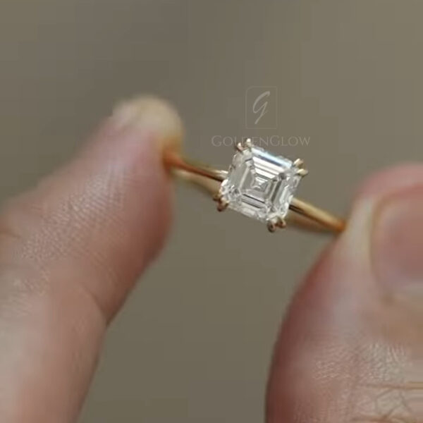 Close-up view of a delicate yellow gold solitaire ring held between fingertips, featuring a square emerald-cut diamond secured by four fine claw prongs. The gemstone displays crisp step facets that reflect light softly, emphasizing clarity and symmetry. The slim polished band curves smoothly around the finger grip, creating a minimal, elegant silhouette. Neutral background tones keep focus on the diamond’s geometry, craftsmanship, and refined proportions.