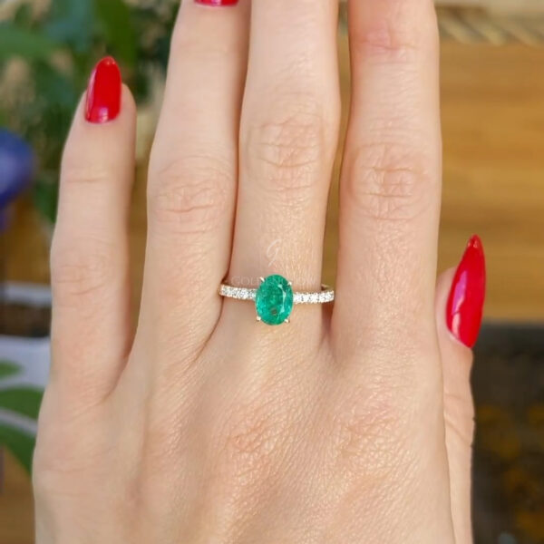 Hand wearing an oval green emerald engagement ring with a slim diamond-accented band, photographed outdoors against lush green leaves. The vivid emerald center stone sits horizontally on the finger, contrasting beautifully with glossy red manicured nails and natural sunlight highlighting the gemstone’s rich color and sparkle.
