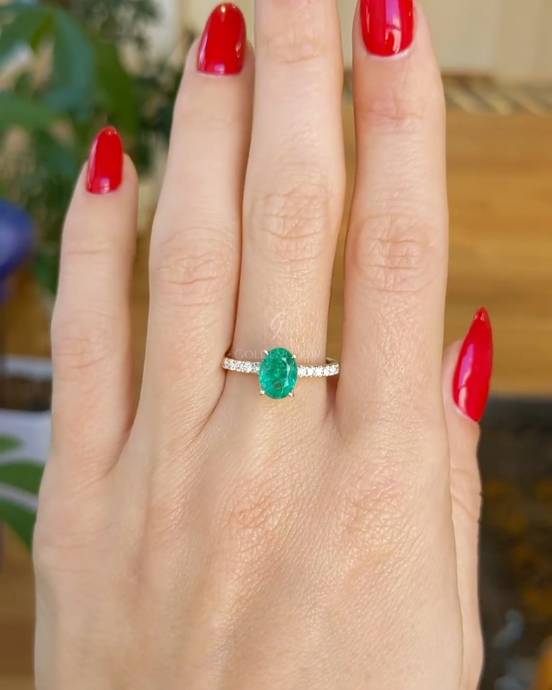 Hand wearing an oval green emerald engagement ring with a slim diamond-accented band, photographed outdoors against lush green leaves. The vivid emerald center stone sits horizontally on the finger, contrasting beautifully with glossy red manicured nails and natural sunlight highlighting the gemstone’s rich color and sparkle.