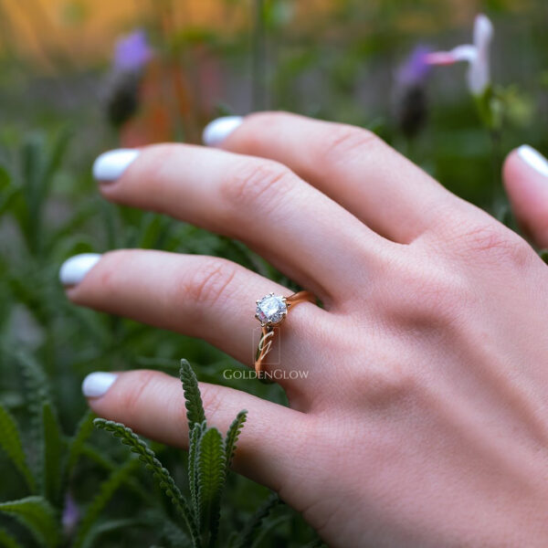 A close-up of a hand wearing a classic solitaire engagement ring with a round-cut moissanite or lab grown diamond set on a slim yellow gold band. The ring is photographed outdoors among green foliage, with soft natural light enhancing the diamond’s sparkle and brilliance. White manicured nails and a blurred floral background create a fresh, romantic, and elegant aesthetic, highlighting the timeless beauty of the ring design.