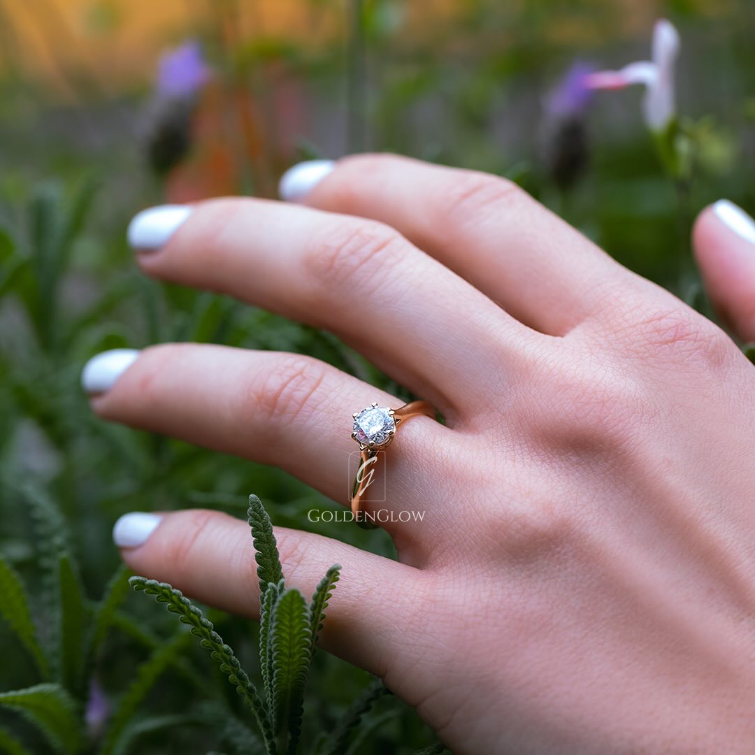 A close-up of a hand wearing a classic solitaire engagement ring with a round-cut moissanite or lab grown diamond set on a slim yellow gold band. The ring is photographed outdoors among green foliage, with soft natural light enhancing the diamond’s sparkle and brilliance. White manicured nails and a blurred floral background create a fresh, romantic, and elegant aesthetic, highlighting the timeless beauty of the ring design.
