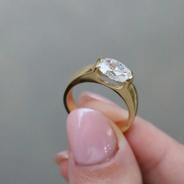 Close-up lifestyle photo of a yellow gold ring worn on a hand. The ring features an oval-cut diamond set horizontally in a smooth, modern bezel-style setting. The polished gold band is wide and sleek, giving a bold yet minimalist look. Soft natural lighting highlights the diamond’s sparkle and the warm tone of the gold. Other delicate gold rings are visible on nearby fingers, with a softly blurred background that keeps focus on the centerpiece ring.
