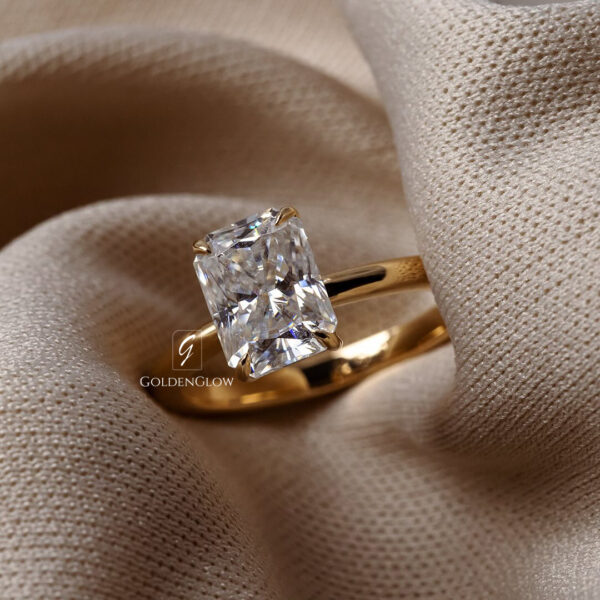 Outdoor lifestyle shot of a woman’s hand wearing a radiant cut moissanite or diamond engagement ring on a slim yellow gold band. The solitaire stone sparkles in natural sunlight, highlighting clean facets and elegant craftsmanship. Soft greenery in the background adds a romantic, modern, and engagement ring aesthetic.