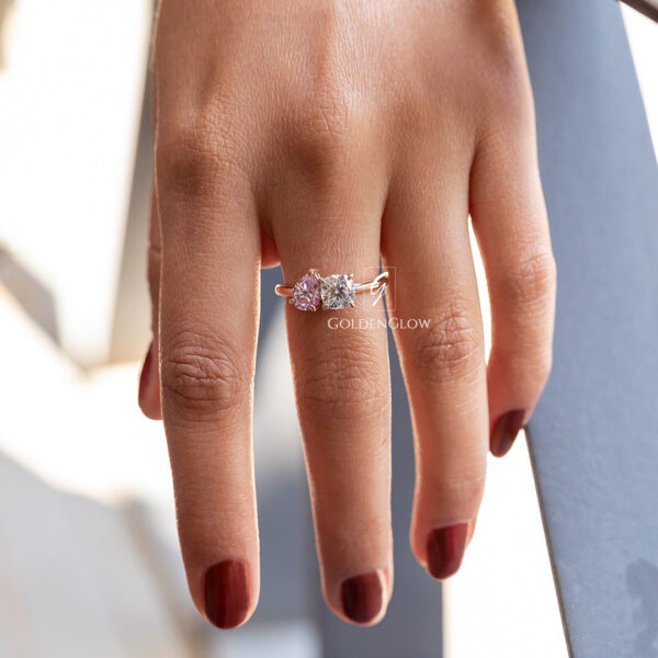 Close-up lifestyle image of a hand wearing a delicate yellow gold ring with two heart-shaped gemstones set side by side. One heart-shaped diamond appears clear and brilliant, while the other has a soft pink hue, creating a romantic and unique contrast. The slim polished band sits gracefully on the finger, complementing natural skin texture and deep red nail polish. Shot in natural light against a softly blurred background, emphasizing fine craftsmanship, elegance, and modern luxury jewelry design.