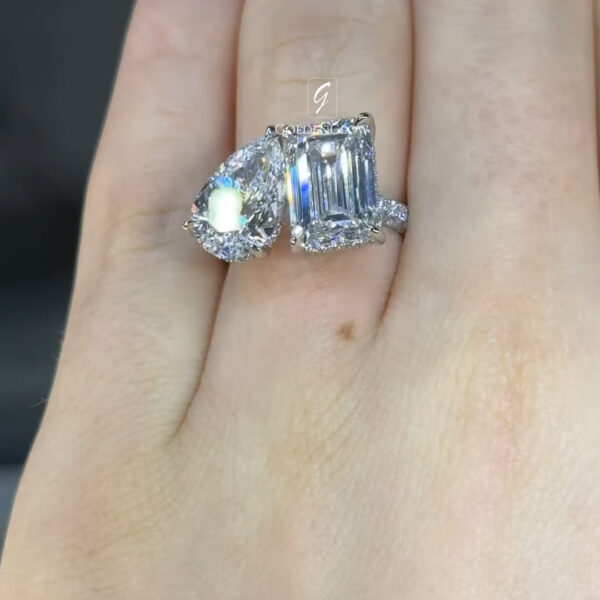 Close-up comparison image of two diamond rings worn side by side on a finger. On the left is an oval-cut diamond ring with brilliant facets and a halo setting that enhances sparkle and fire. On the right is an emerald-cut diamond ring with elongated step-cut facets, set in a prong setting with small accent diamonds along the band. Both rings are white metal and reflect bright light. The background is softly blurred, and a subtle GOLDENGLOW watermark appears above the diamonds.