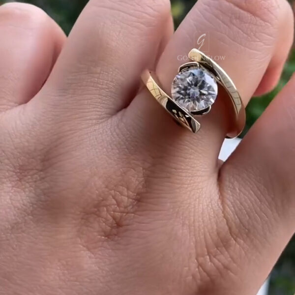 A detailed close-up of a hand wearing a contemporary yellow gold ring with a round brilliant-cut lab-grown diamond at the center. The diamond is held in a smooth bezel setting and framed by an elegant bypass-style band that curves around the stone from both sides. The polished gold surface features subtle engraved detailing and reflects natural light, while the diamond displays strong sparkle and clarity. The background is softly blurred greenery, highlighting the ring’s modern, minimalist design.