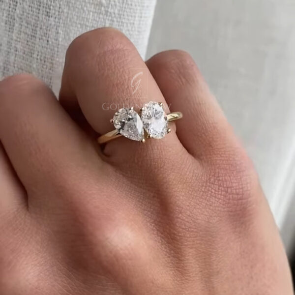Close-up of a hand wearing a delicate two-stone diamond ring on the finger. The ring features two clear, pear-shaped diamonds set side by side in a yellow gold band, each secured with fine prongs. The gemstones reflect soft natural light, creating subtle sparkle rather than sharp flashes. The hand is gently curved, showing natural skin texture and fine lines. A light, textured fabric curtain in the background adds a calm, airy feel and highlights the elegance and simplicity of the ring’s design.