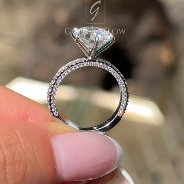 Close-up side profile of a diamond engagement ring held between the thumb and index finger. The ring features a round brilliant-cut center stone secured in an elevated four-prong setting, allowing light to pass through the pavilion. The white metal band is lined with small pavé diamonds along the shoulders, adding continuous sparkle. The polished band reflects sunlight,