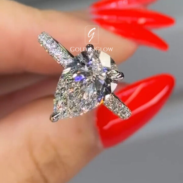 Close-up angled view of a diamond engagement ring held between the thumb and index finger. The ring features a pear-shaped clear center stone secured by prongs, set on a white metal band with pavé diamonds along the shoulders. The center stone shows bright sparkle and colorful light reflections across its facets. The fingers have long, glossy red nails that contrast with the silver-toned metal. A smooth gray background keeps focus on the ring’s elegant shape, brilliance, and detailed craftsmanship.