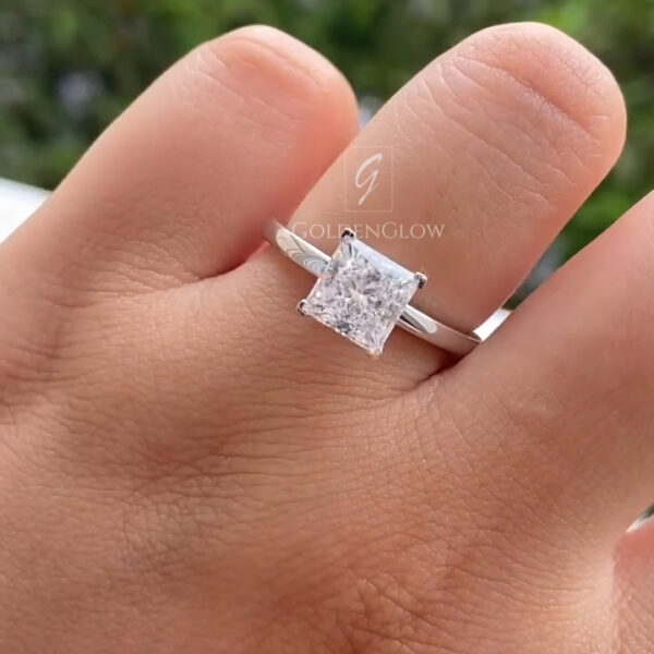 Close-up of a hand wearing a solitaire engagement ring with a square princess-cut clear center stone set in four prongs. The diamond sits on a slim, polished white metal band, emphasizing a clean, modern design. Soft lighting highlights the stone’s sharp corners, internal sparkle, and symmetrical shape against a dark background.