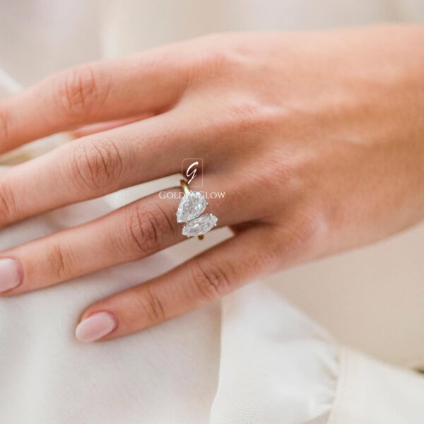 A close-up of a hand resting on soft white fabric, wearing a delicate engagement ring with two pear-cut moissanite or diamonds. The stones are arranged in a vertical, modern design on a slim yellow-gold band. The diamonds reflect gentle natural light, highlighting their brilliance and clarity. The hand features natural nude-pink manicured nails, and the light, neutral background enhances the elegant, minimal, and timeless appeal of the ring.
