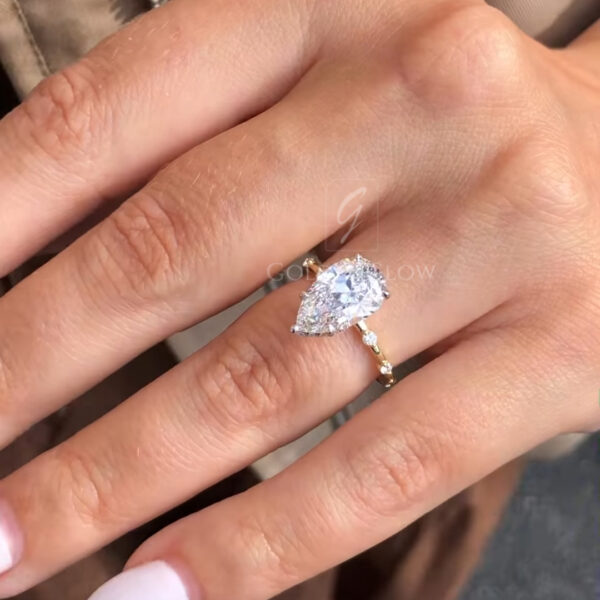 Close-up of a pear-shaped diamond engagement ring worn on a hand, featuring a brilliant teardrop center stone set in yellow gold. The ring sparkles in natural light, with a slim band and delicate accent diamonds, photographed outdoors against a neutral background for scale and realism.