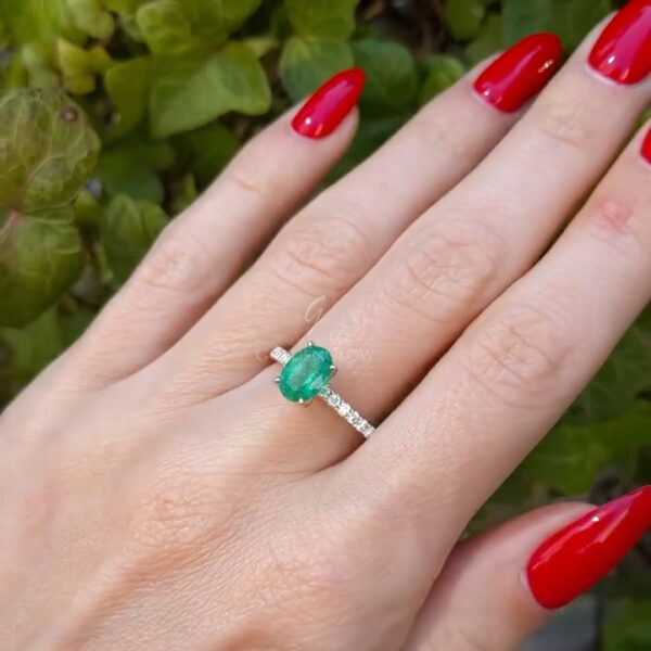 Close-up of a hand displaying an oval emerald ring with a pavé diamond band, photographed indoors with warm lighting. The emerald gemstone is centered on the finger, framed by bright red nail polish and a softly blurred background that emphasizes the fine jewelry craftsmanship and elegant proportions.