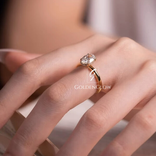 A close-up shot of a hand wearing a solitaire engagement ring with an oval-cut moissanite or lab grown diamond. The gemstone is held in a classic four-prong yellow-gold setting, paired with a slim, smooth, and polished band. The stone reflects soft natural light, highlighting its brilliance and clean facets. The ring is styled on elegant fingers with natural nude-pink manicured nails. A softly blurred, warm-toned background enhances the luxurious.