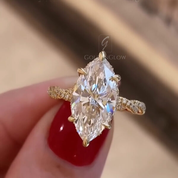 Close up view of a marquise cut diamond engagement ring held between fingers with red polished nails. The elongated diamond is set vertically in yellow gold prongs, showing sharp facets and bright reflections. The twisted yellow gold band features pavé diamonds along each shoulder, adding texture and sparkle. Soft indoor lighting highlights the stone clarity and metal finish, while a blurred neutral background keeps attention focused on the ring’s design.