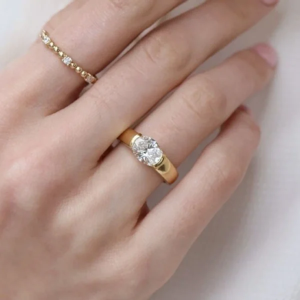 Close-up of a yellow gold ring held between fingers with glossy nude-pink manicured nails. The ring features an oval-cut diamond set horizontally in a sleek, modern bezel-style setting. The polished gold band is smooth and slightly tapered, giving a minimalist, contemporary look. A soft gray background keeps focus on the diamond’s sparkle and the clean lines of the gold setting.