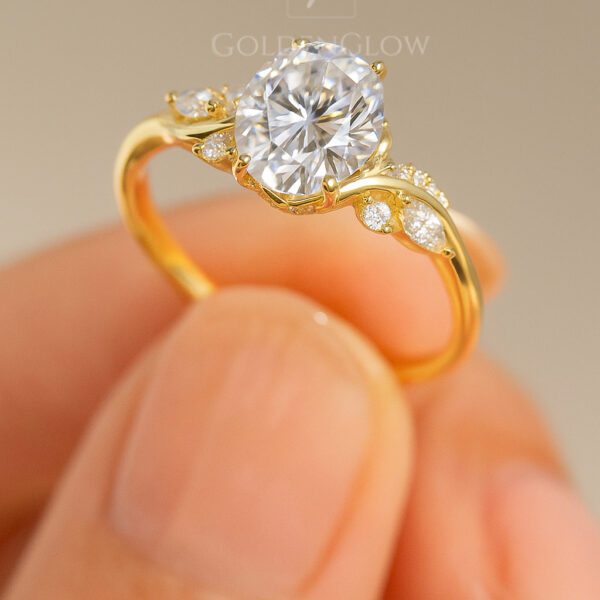 Close-up side view of a yellow gold engagement ring held between fingers, featuring a round brilliant center stone secured in claw prongs. The sculpted band curves gracefully and is accented with small pavé diamonds along the shoulders, highlighting the elegant setting and refined craftsmanship against a soft neutral background.