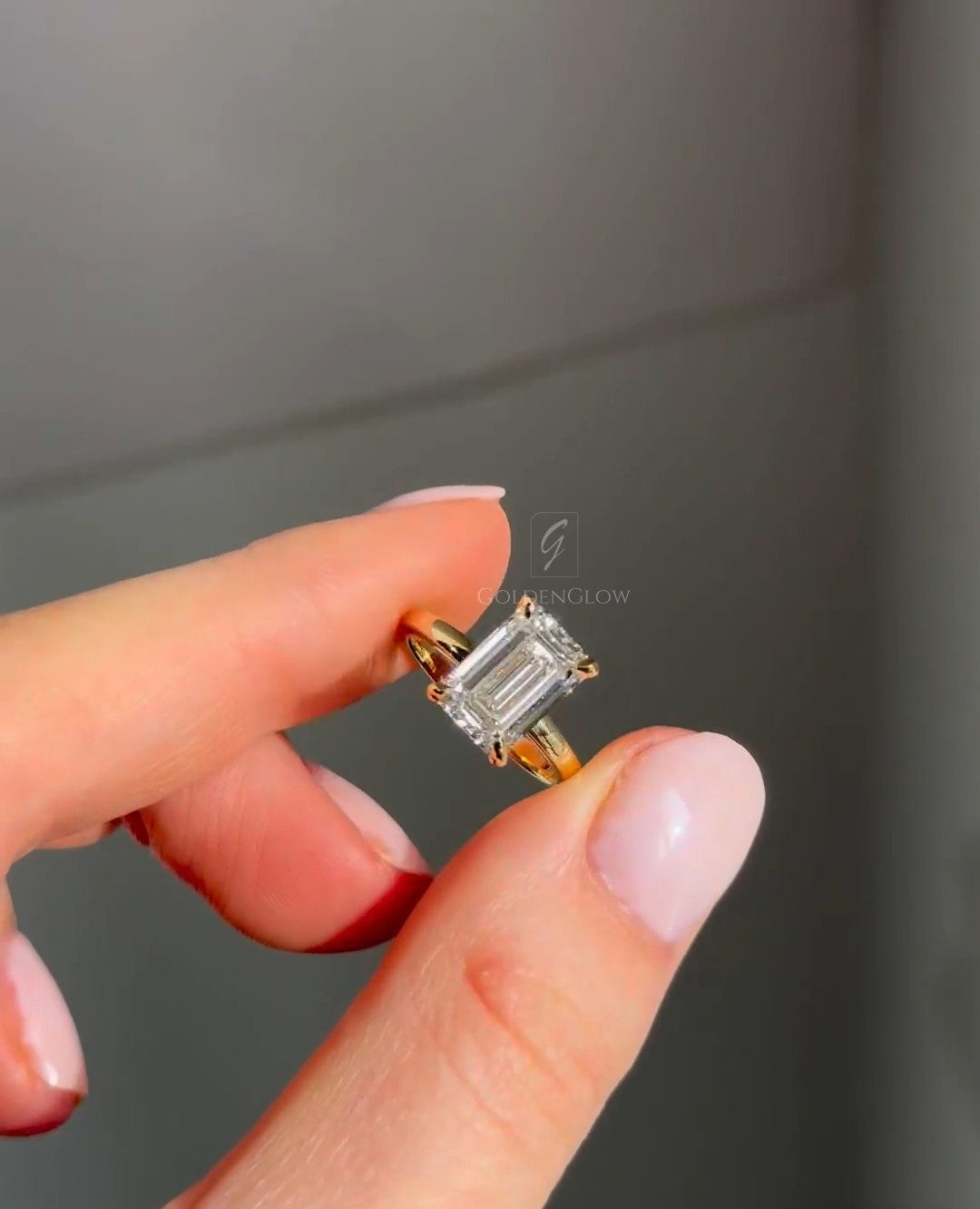 Close-up of an emerald-cut diamond engagement ring held between fingers, featuring a rectangular step-cut stone set in a yellow gold band with four prongs. The diamond’s clean lines and mirror-like facets reflect soft natural light against a neutral background, highlighting the elegant proportions, clarity, and minimalist solitaire design suitable for modern and jewelry lovers.